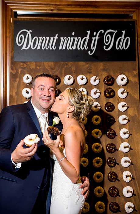 Bride kissing the grooms cheek while standing in front of a donut wall holding donuts.