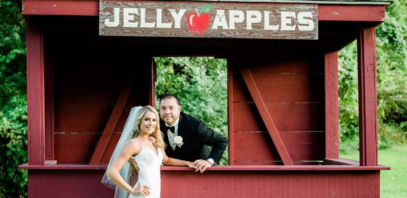 Bride and groom pose in front of a Jelly Apple stand.