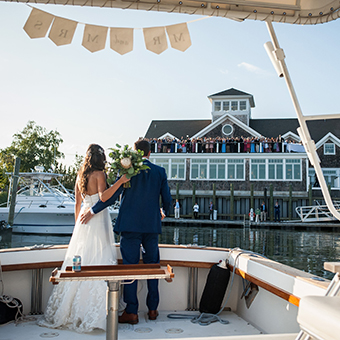 Bride and groom pose on a boat looking towards the wedding party at the venue.