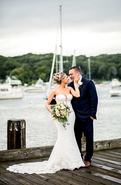 Bride holding grooms face while standing in front of a harbor.