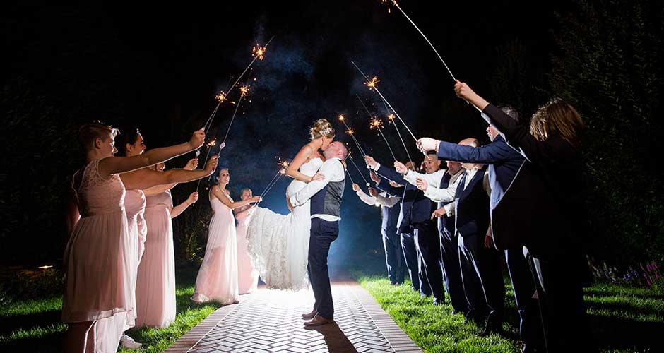 Grooms lifts the bride in the middle of the wedding party holding sparklers.