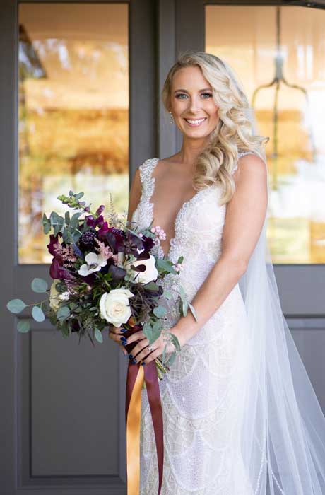 Bride standing holding her purple and white bouquet in front of glass wooden doors.