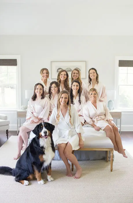 Bride is sitting with her bridesmaids and her dog on a bed.