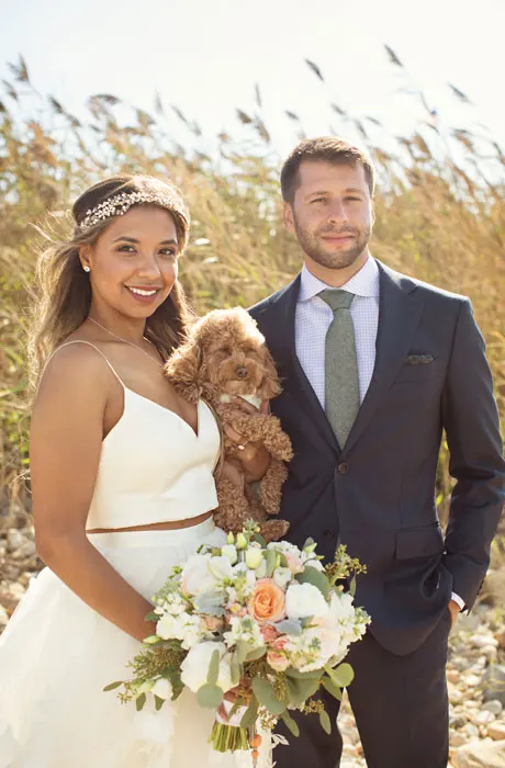 Bride and groom are holding their dog in a field.