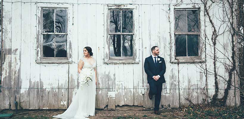 Bride and Groom standing separately next to a vintage white barn.