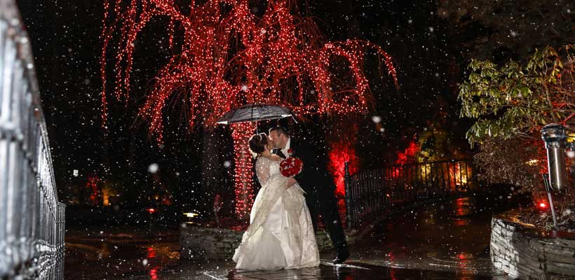 Bride and groom kiss under red holiday lights during snowfall.