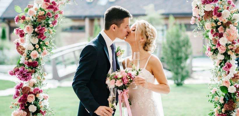 Bride and groom kiss outdoors under a flower arch.