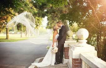 A bride and groom kissing on a bridge while the brides veil is blowing in the wind.